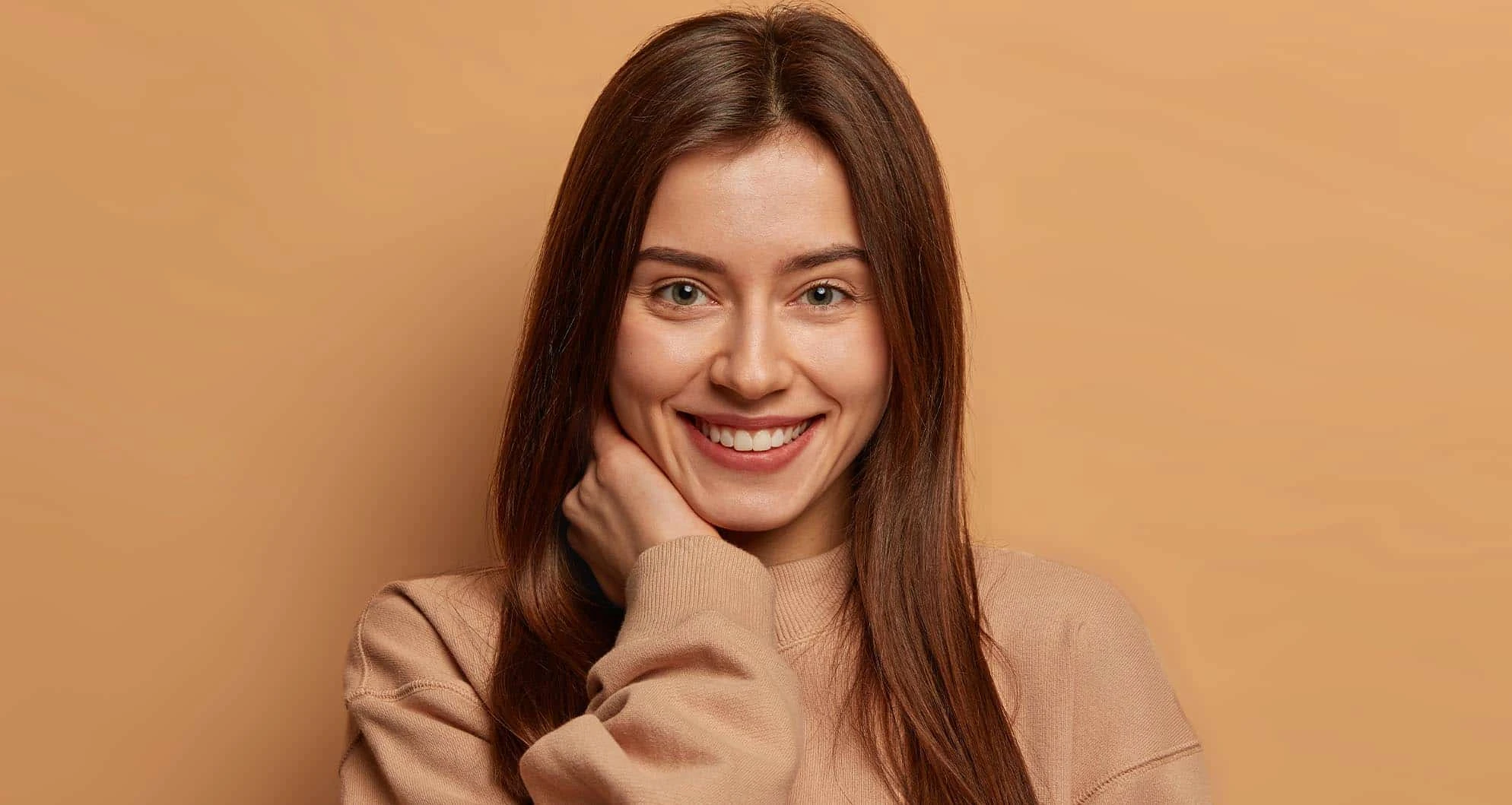 A young woman with long brown hair smiling warmly against a plain tan background, wearing a beige sweater. Her natural, glowing look conveys confidence and radiance, making it ideal for promoting Med Spa Black Friday specials at Inspire Medical Spa in South Jordon, Utah.