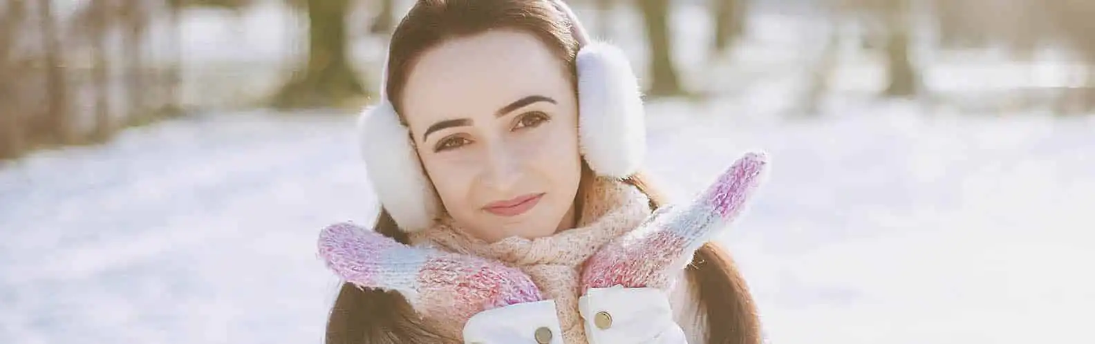 A woman bundled in earmuffs, scarf, and gloves smiles in a snowy outdoor setting, highlighting the importance of Winter Skincare Essentials at Inspire Medical Spa in South Jordan, Utah.
